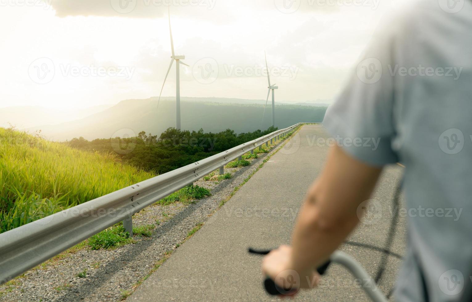 Selective focus on wind farm with rearview of person riding a bicycle as foreground. Wind energy. Wind power. Sustainable, renewable energy. Wind turbines generate electricity. Sustainable lifestyle. photo