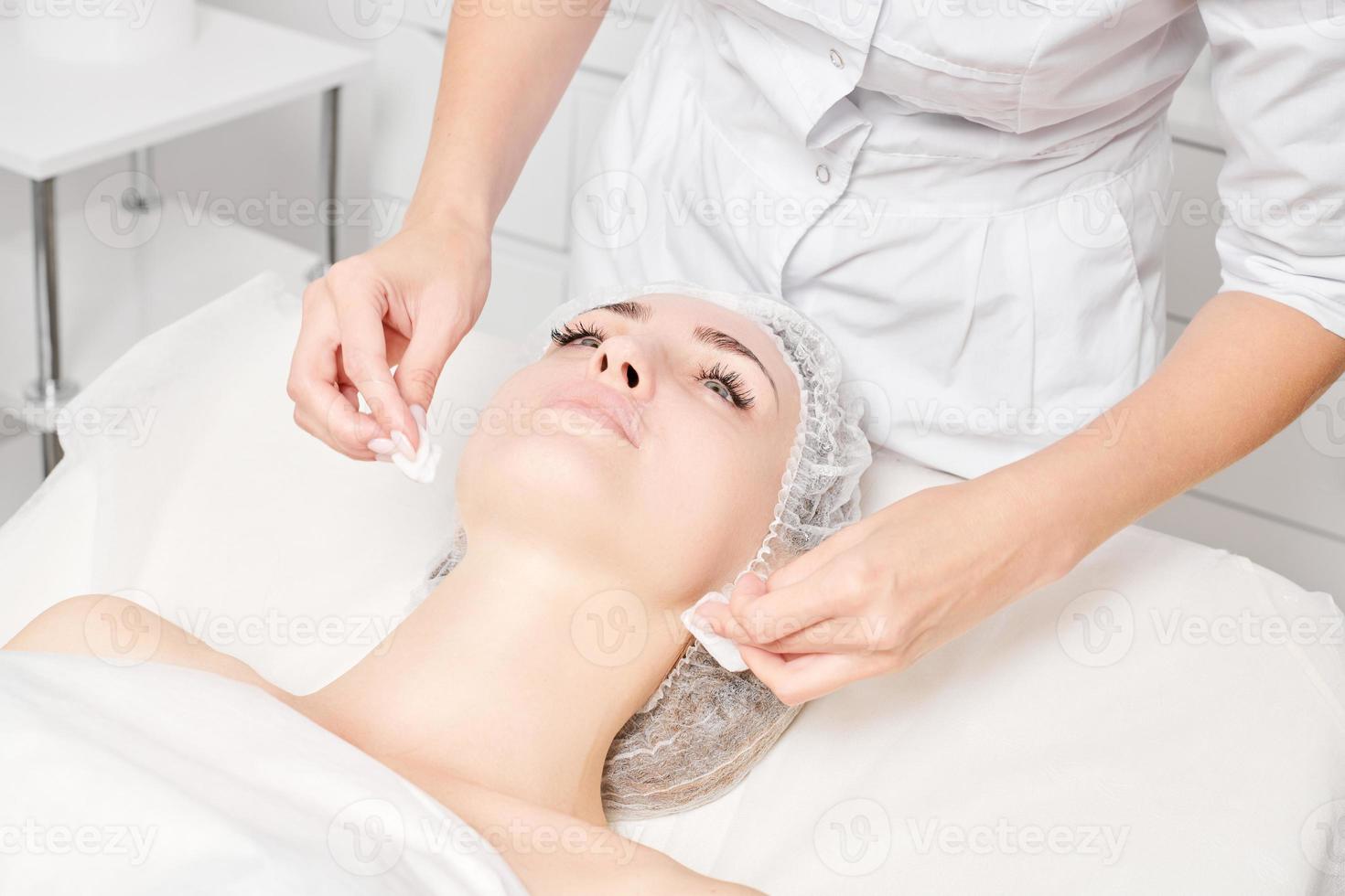 Cosmetologist removing facial cream mask from woman face skin with cosmetic wipes in beauty salon photo