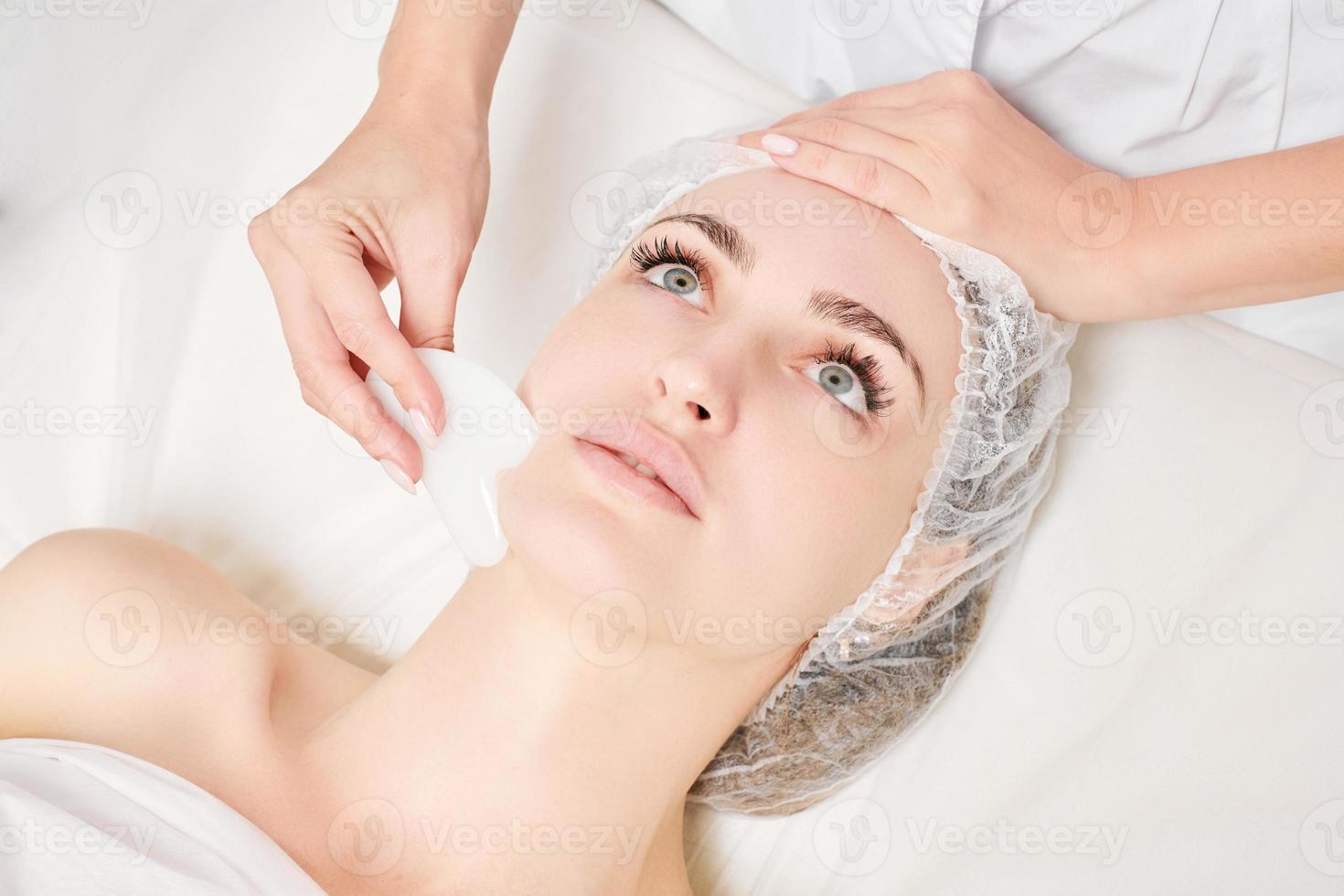esteticista haciendo masaje facial con piedra gua sha de mujer piel facial para drenaje linfático foto