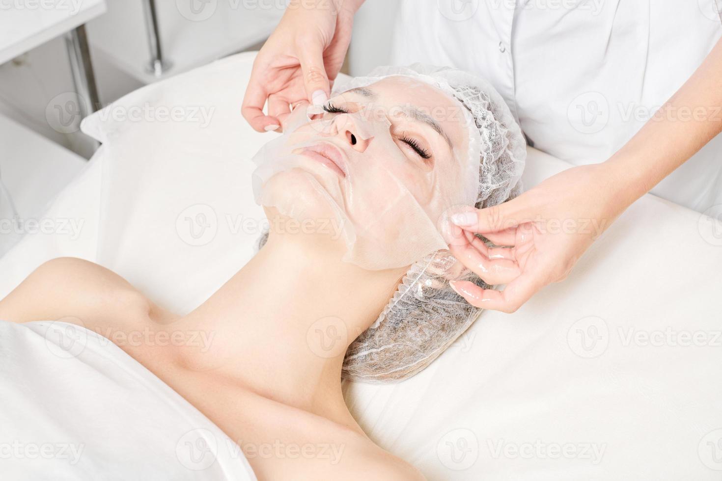 esteticista suaviza la máscara de hoja en la cara de la mujer para rehidratar la piel de la cara, procedimiento en el salón de belleza foto