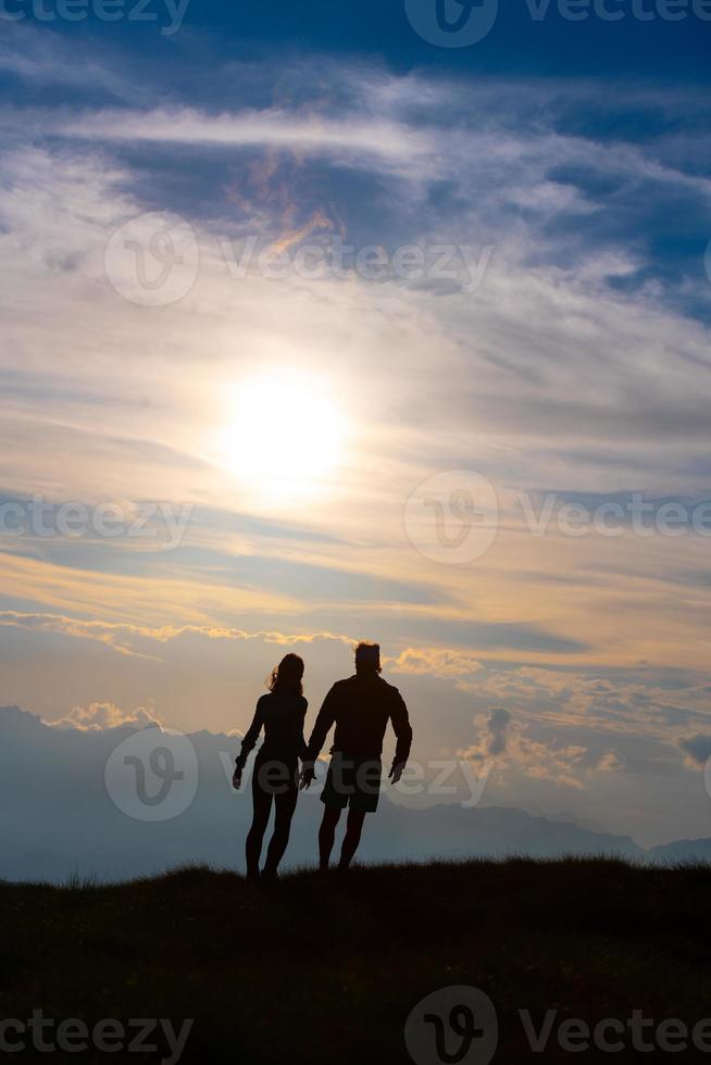 pareja en silueta tomados de la mano caminando hacia nubes coloridas al atardecer en las montañas foto