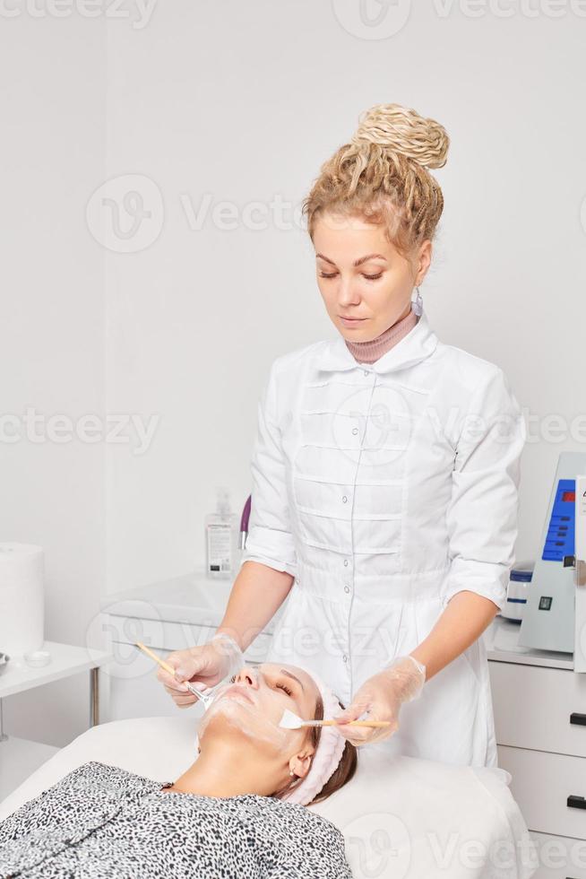 Cosmetologist applying cosmetic cream mask on female face for rejuvenation procedure in beauty salon photo