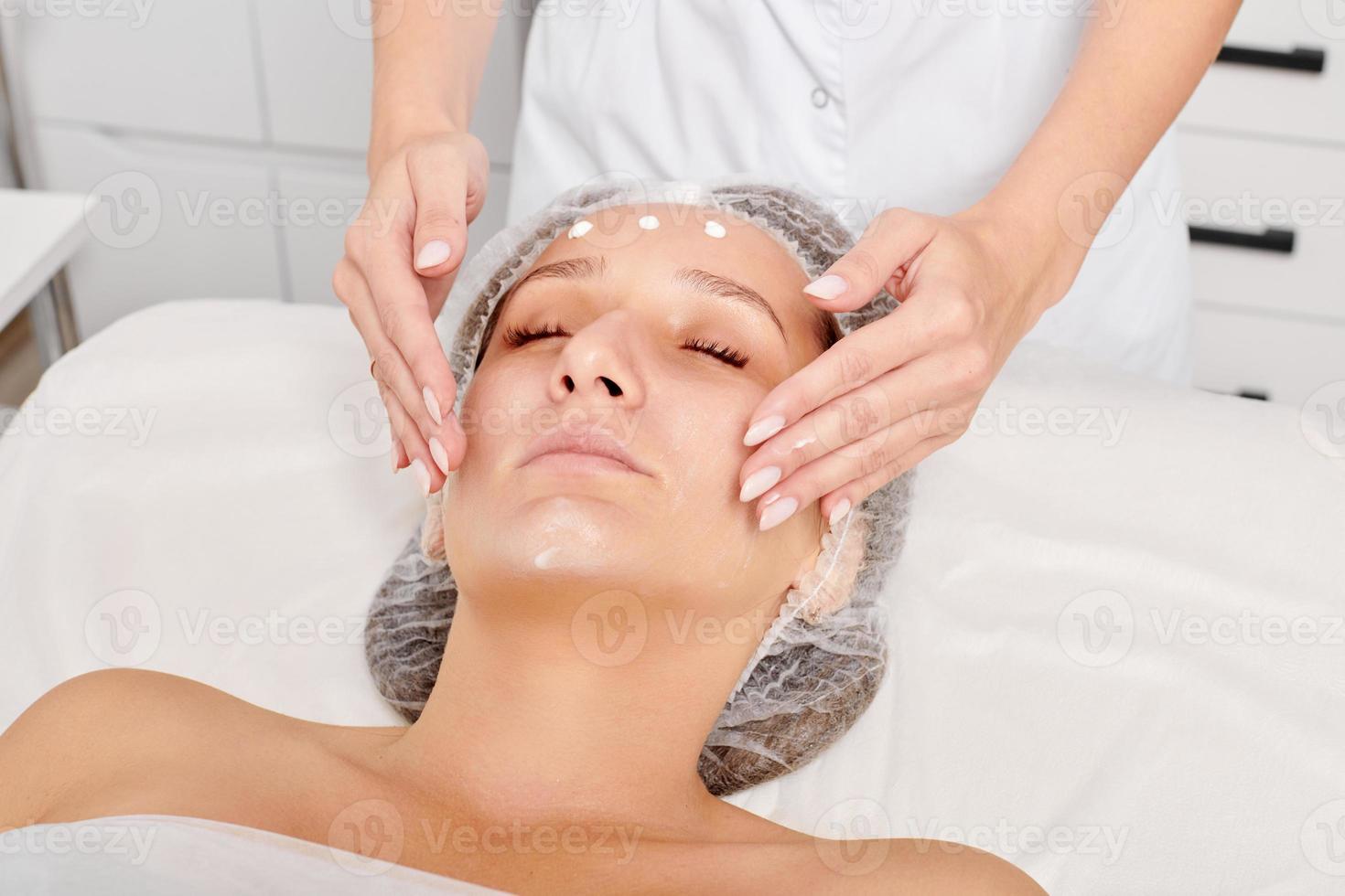 Beautician rubbing moisturizing cream mask into woman face skin for rejuvenation in beauty salon photo