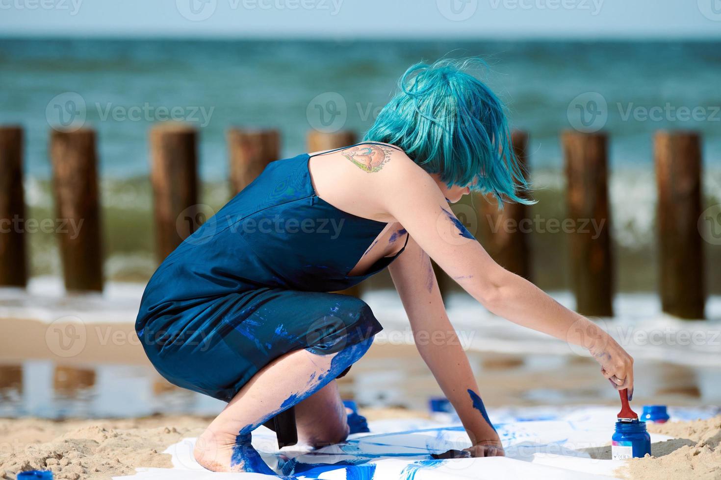 Talented blue-haired woman performance artist smeared with gouache paints on large canvas on beach photo