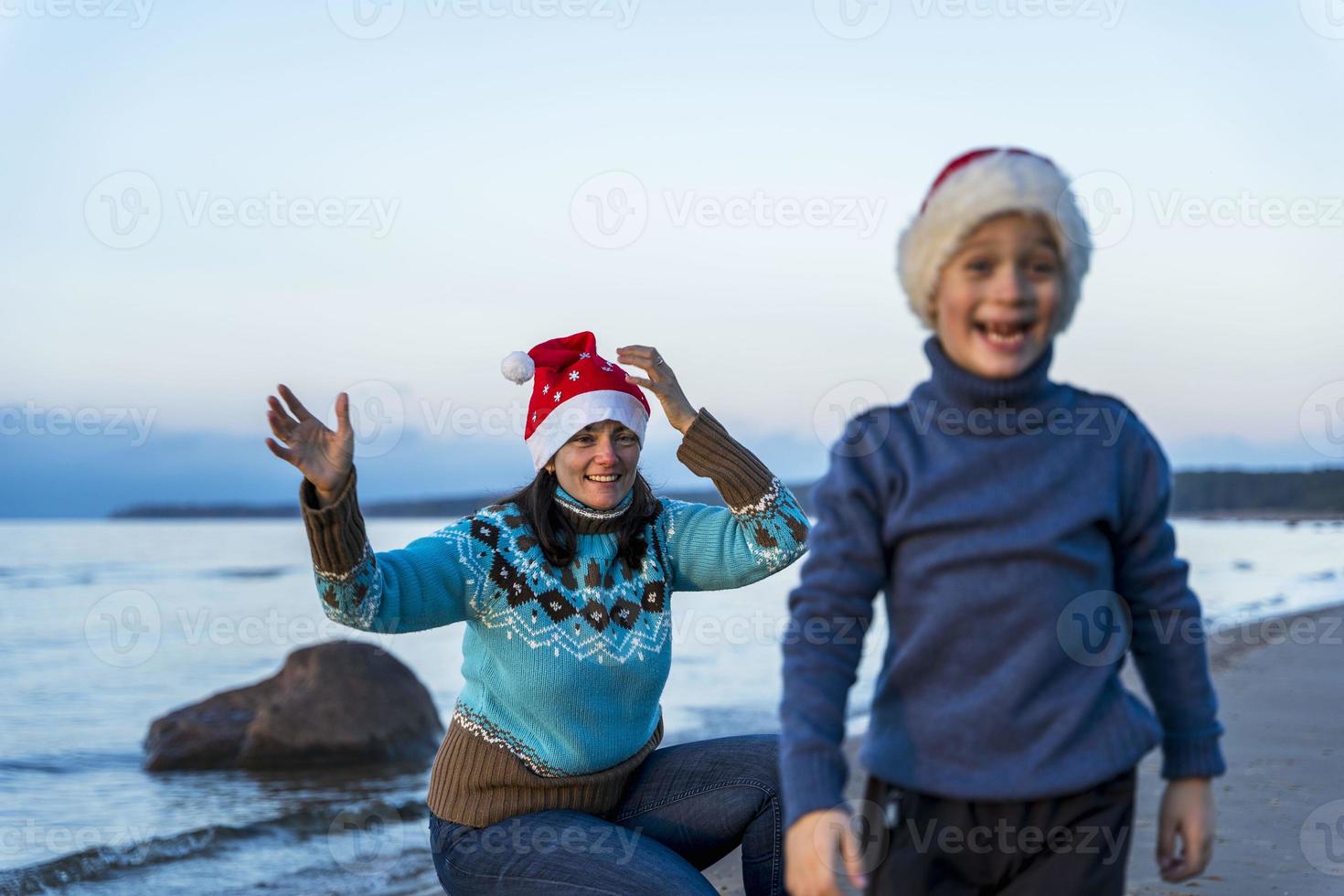 Happy mom and little son celebrate Christmas on the beach in Christmas hats, the boy laughs merrily, blurr photo