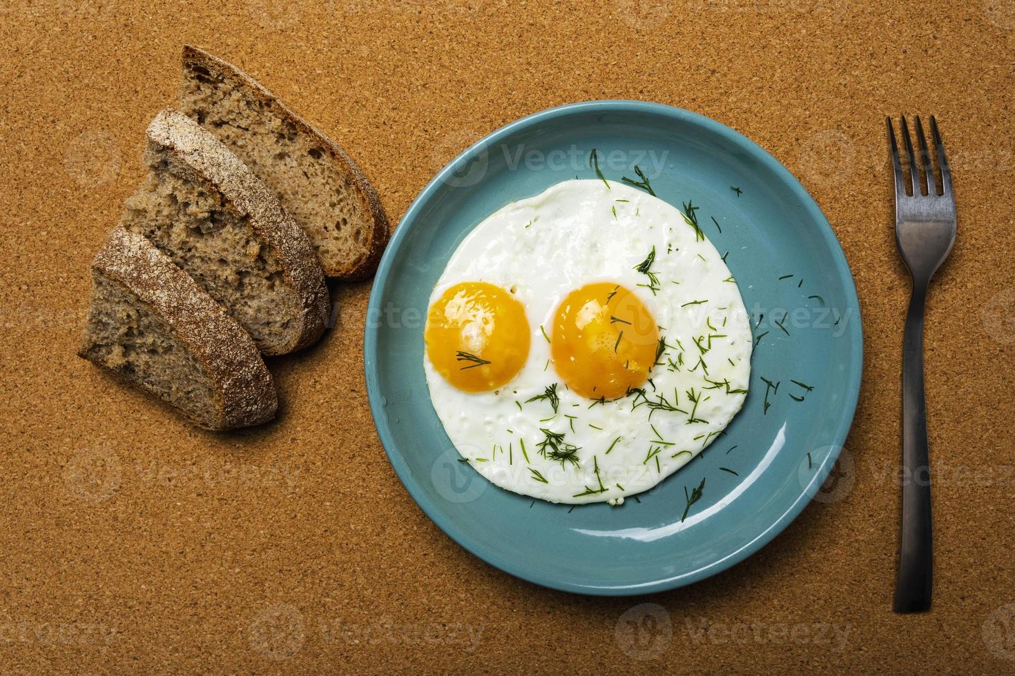 fried eggs on a turquoise plate sprinkled with dill, fork and pieces of bread, top view, breakfast photo