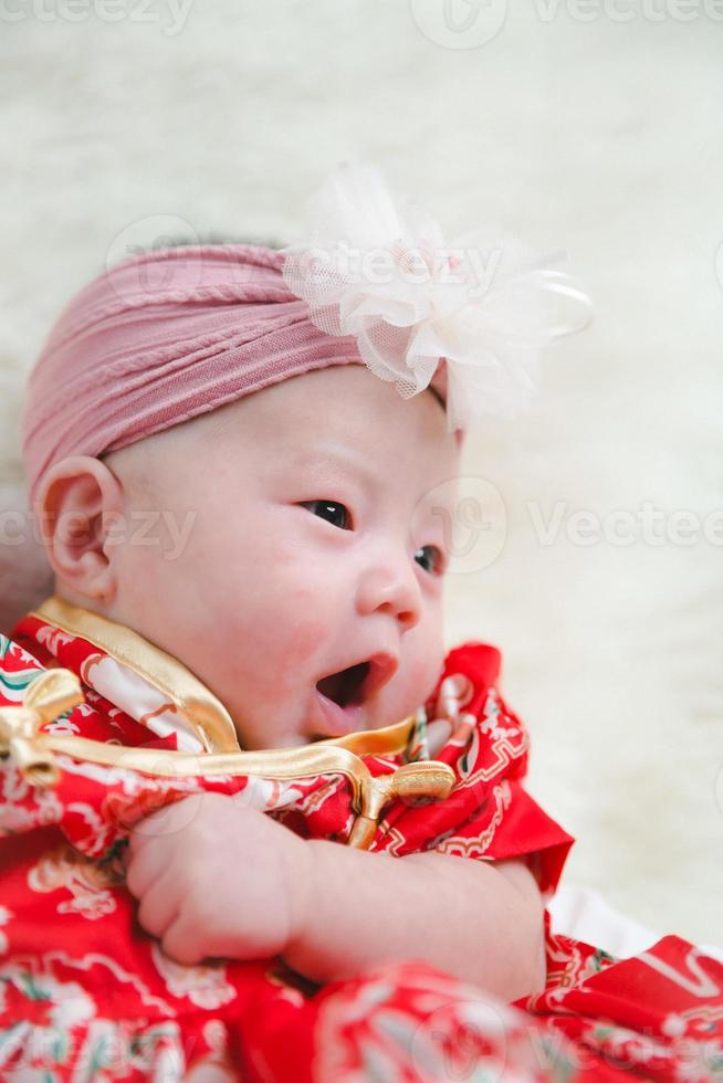 Closeup cute newborn baby in red bodysuit lying down alone on bed. Adorable infant rests on white bedsheets, staring at camera looking peaceful. Infancy, healthcare and paediatrics, babyhood concept. photo