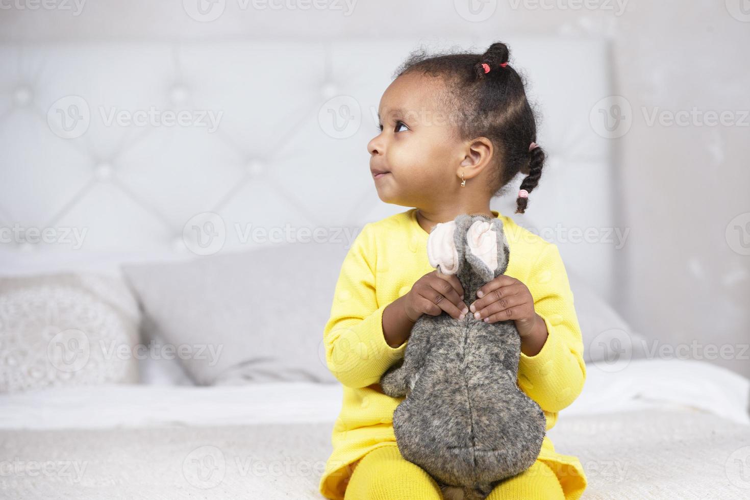 Little African American girl in a yellow dress sits on the bed with a soft toy. photo