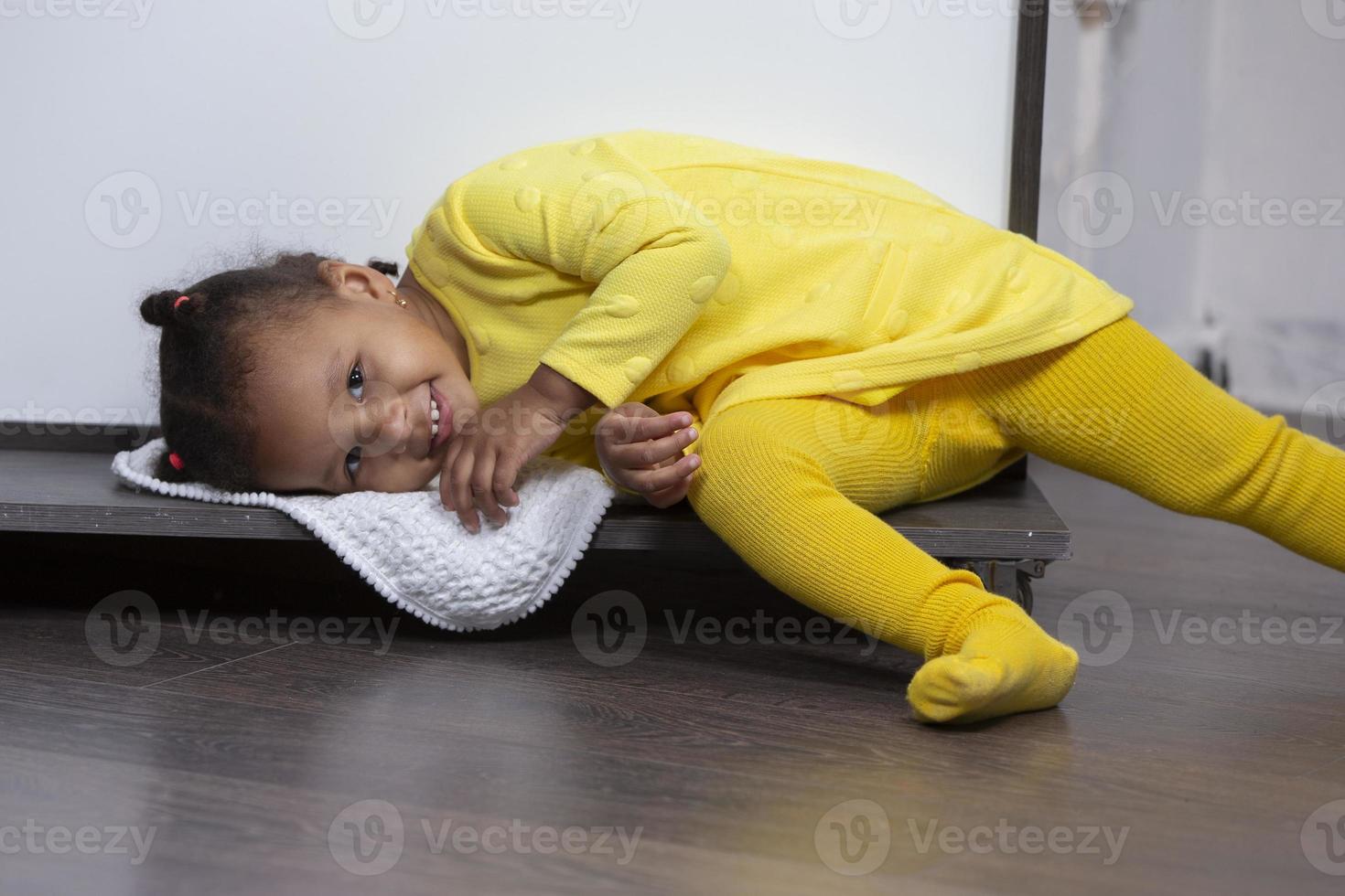 A little African American girl in a yellow dress lies on the floor and smiles. photo