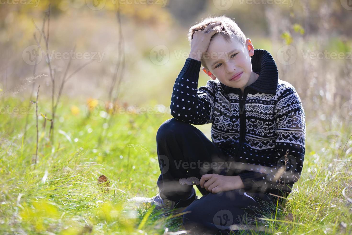 A handsome calm boy in a knitted sweater sits on the grass on the field. photo