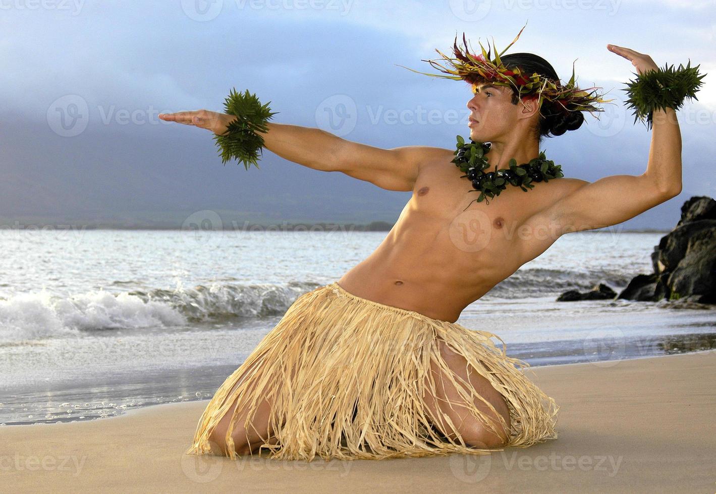 This very handsome male hula dancer is posed in a traditional position on the sand right on the wet sand next to the ocean. photo