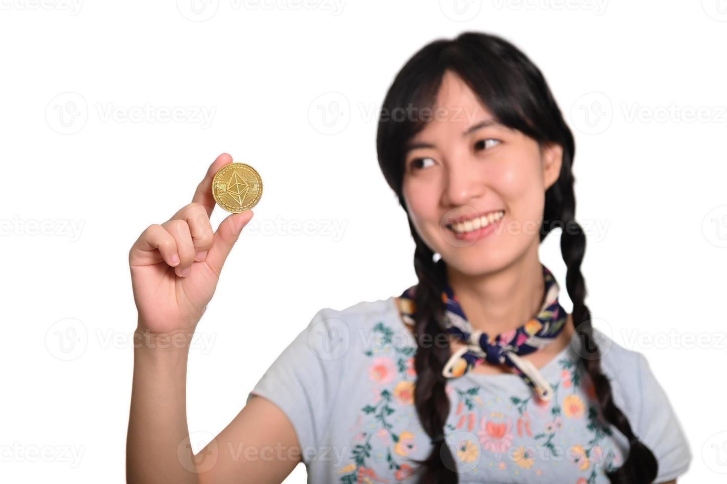 retrato de una hermosa joven asiática feliz vestida de denim sosteniendo una moneda criptográfica sobre fondo blanco. foto