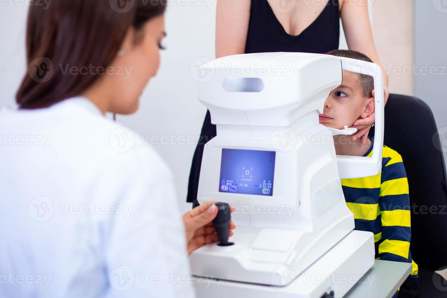 Female oculist using machine for checking eye sight in clinic. Little boy looking at equipment and doctor testing eye pupil in optical store. Concept of eye care and health. photo