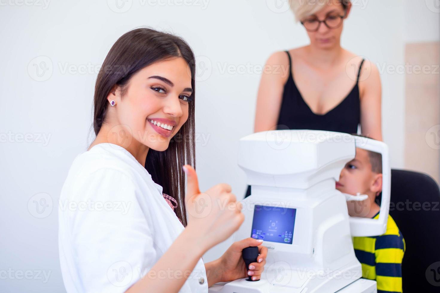 oculista femenina que usa una máquina para controlar la vista en la clínica. niño pequeño que mira el equipo y el médico prueba la pupila del ojo en la tienda óptica. concepto de cuidado ocular y salud. foto