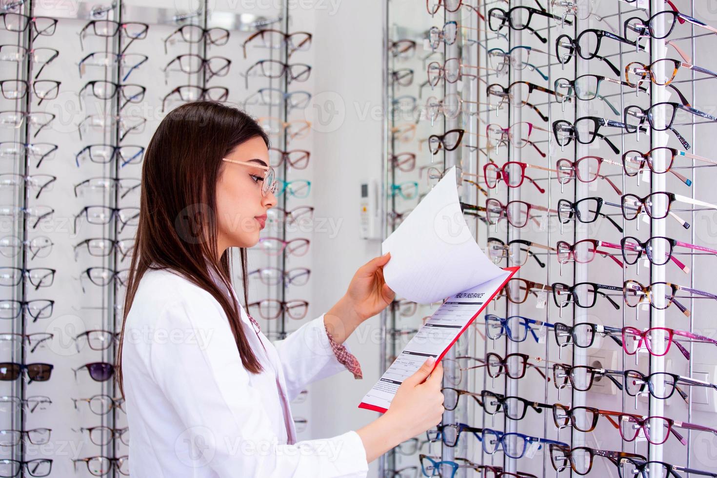 Attractive female ophthalmologist writing data to a clipboard, working in an optical store. Healthcare and medicine concept photo