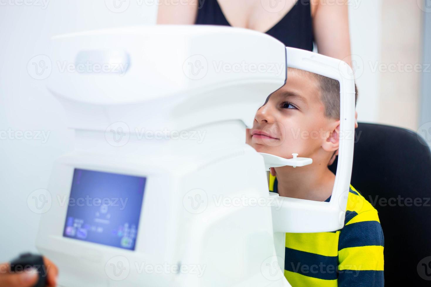 Female oculist using machine for checking eye sight in clinic. Little boy looking at equipment and doctor testing eye pupil in optical store. Concept of eye care and health. photo