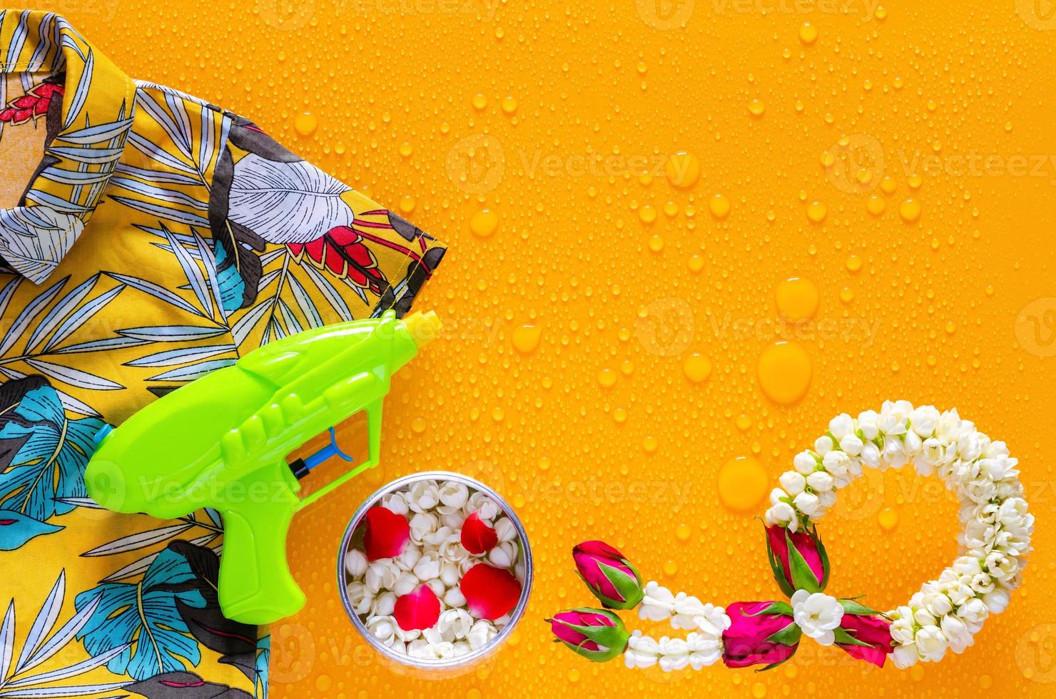 Songkran festival background with colorful shirt, water gun, flowers in water bowl and jasmine garland put on clear mirror that have wet yellow background below. photo
