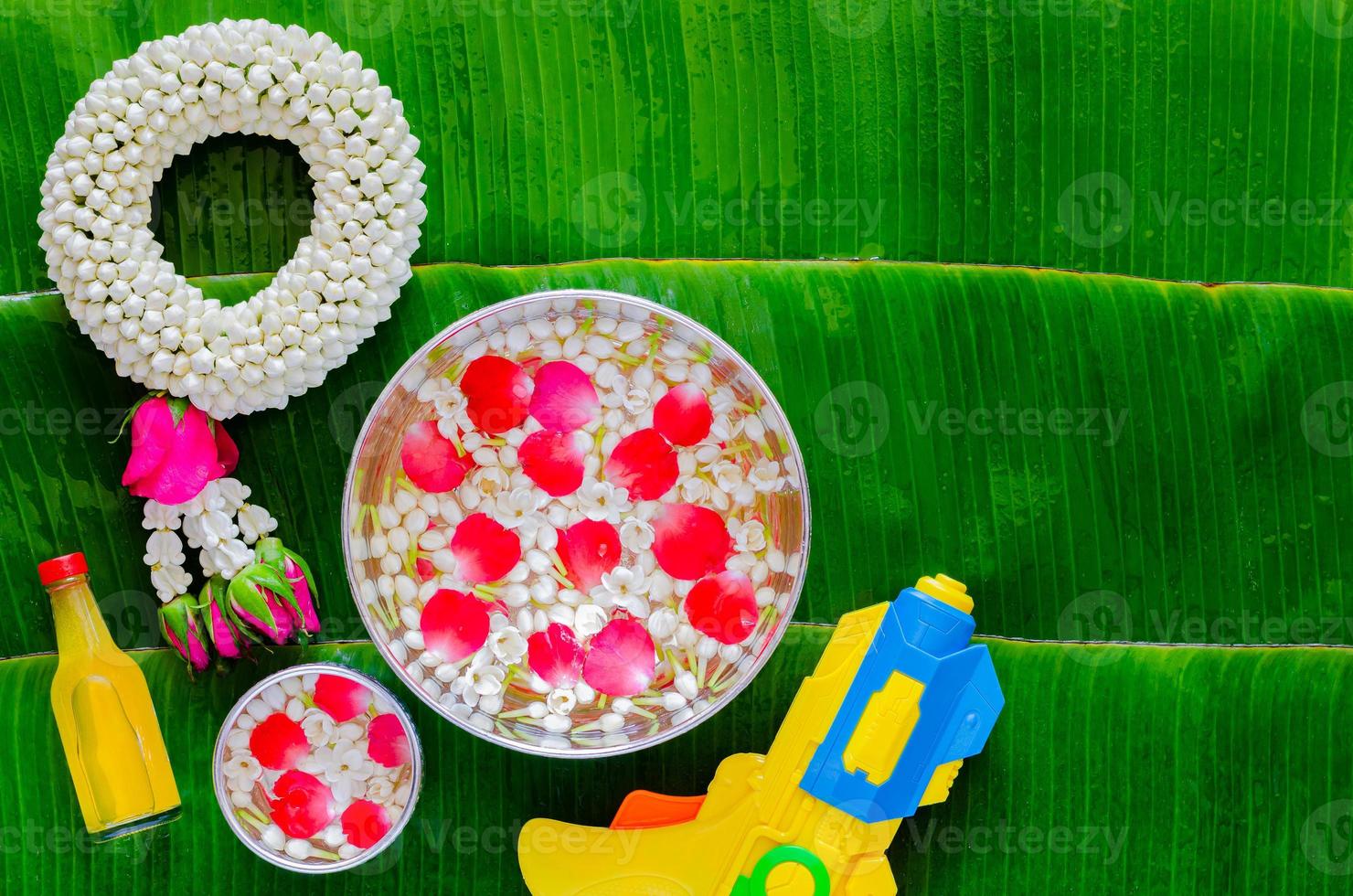 Songkran festival background with jasmine garland, flowers in water bowls, scented water for blessing and water gun on wet banana leaf background. photo