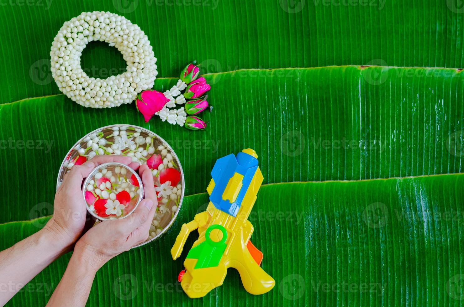 Hand holding water bowl with flowers for blessing that have water gun and jasmine garland put on wet banana leaf background for Songkran festival. photo