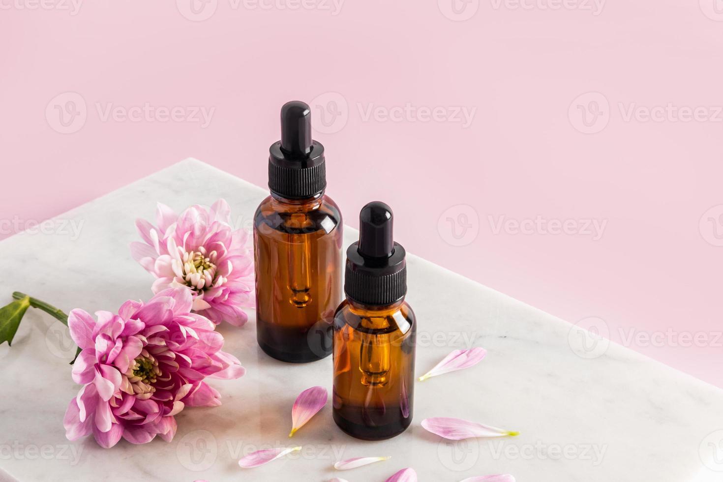 dos botellas cosméticas de vidrio ámbar con una pipeta con un remedio natural para el cuidado de la piel de la cara y el cuerpo sobre un podio de mármol con flores. foto