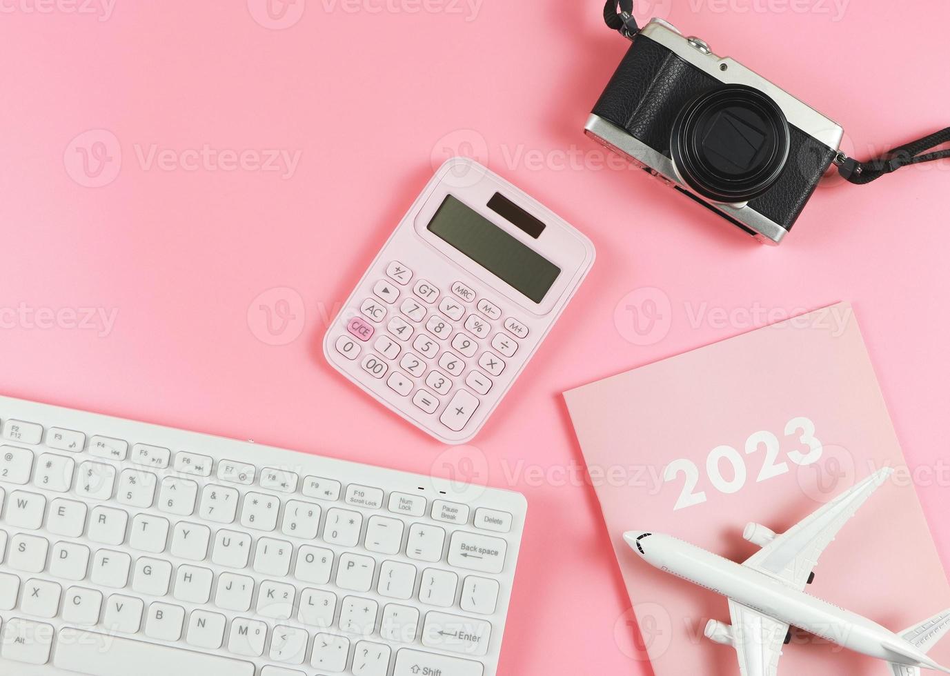 plano de teclado de computadora blanco, diario rosa 2023, modelo de avión, calculadora rosa y cámara sobre fondo rosa con espacio de copia. concepto de planificación de viajes. foto