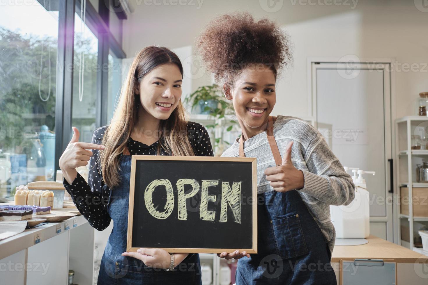 Two young female shopkeepers show open sign board with cheerful smiles in refill store shop, happy work with organic products, zero waste groceries, eco-friendly merchandise, and sustainable business. photo