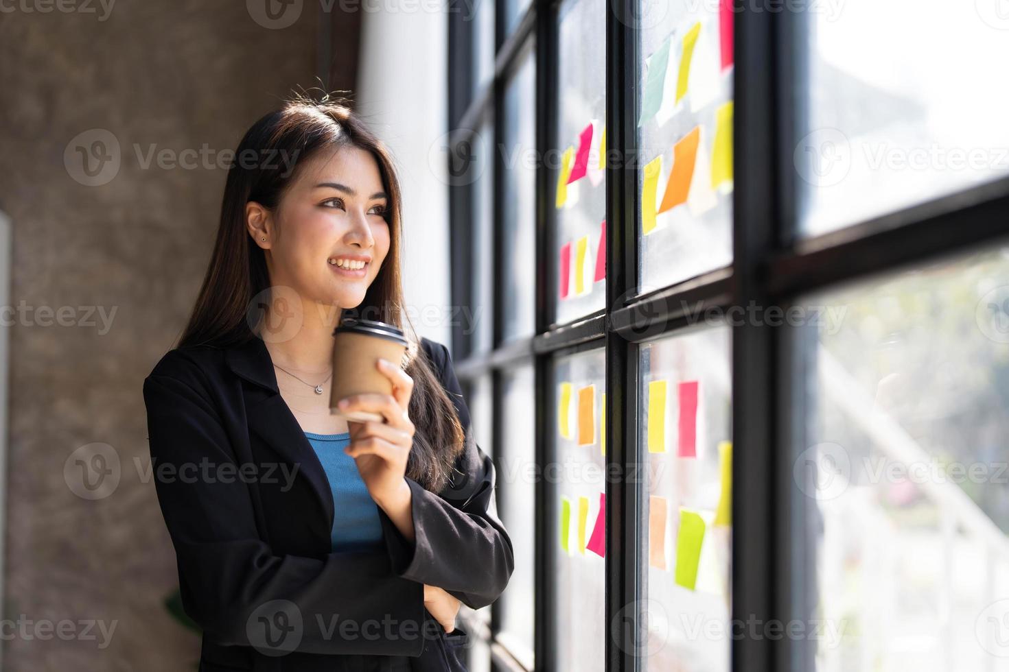 joven empresaria de pie tomando café y pensando en la estrategia de una nueva puesta en marcha mientras mira por la ventana en una oficina moderna. foto