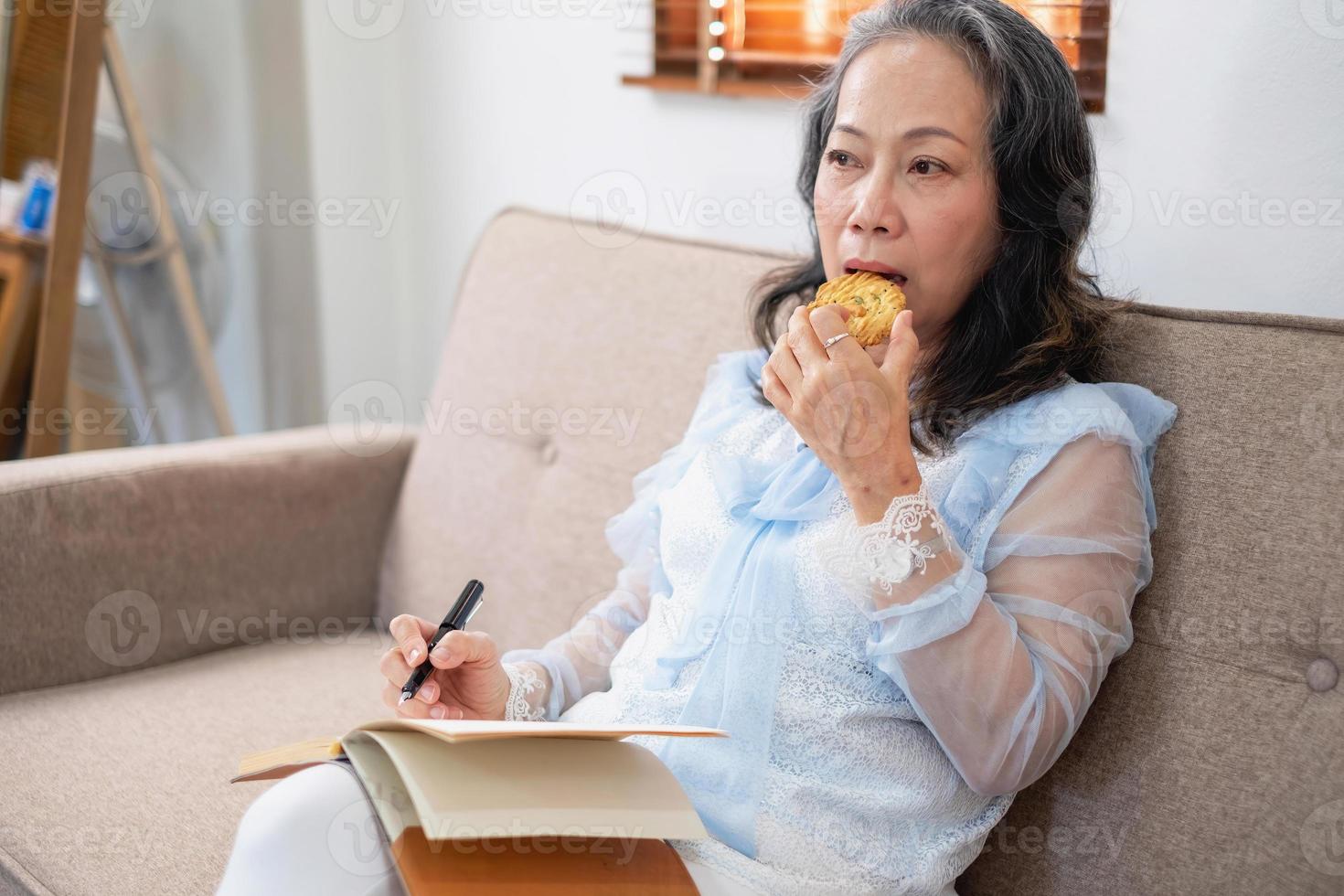 anciana asiática relajándose en el sofá de su casa comiendo galletas en sus manos y escribiendo sus notas en su cuaderno para las vacaciones. concepto de descanso y relajación foto