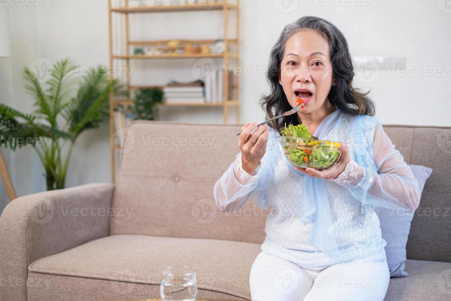 anciana asiática sentada comiendo ensalada de verduras y comida saludable y comiendo felizmente en el sofá de la casa para un cuerpo sano. concepto de comida saludable foto