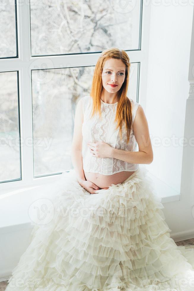 red-haired pregnant young girl in a white dress near the window photo