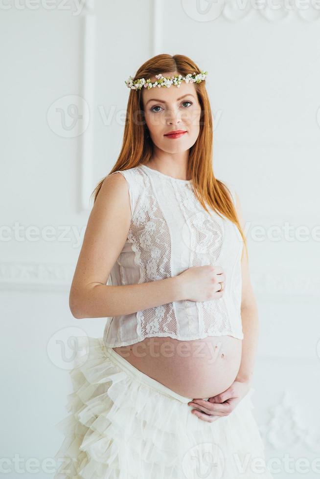 joven embarazada pelirroja con un vestido blanco cerca de la pared blanca foto