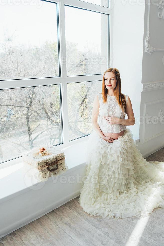 red-haired pregnant young girl in a white dress near the window photo