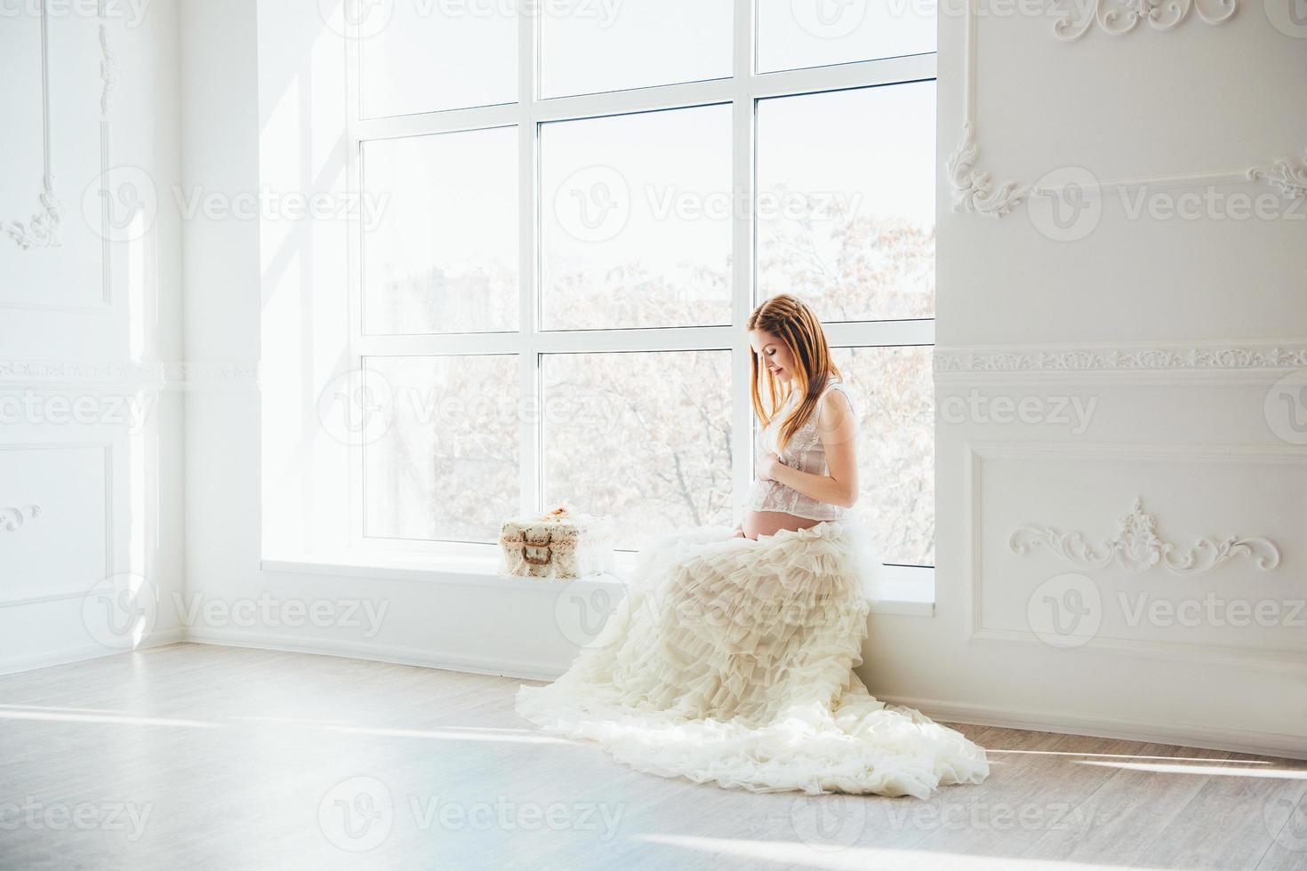 red-haired pregnant young girl in a white dress near the window photo