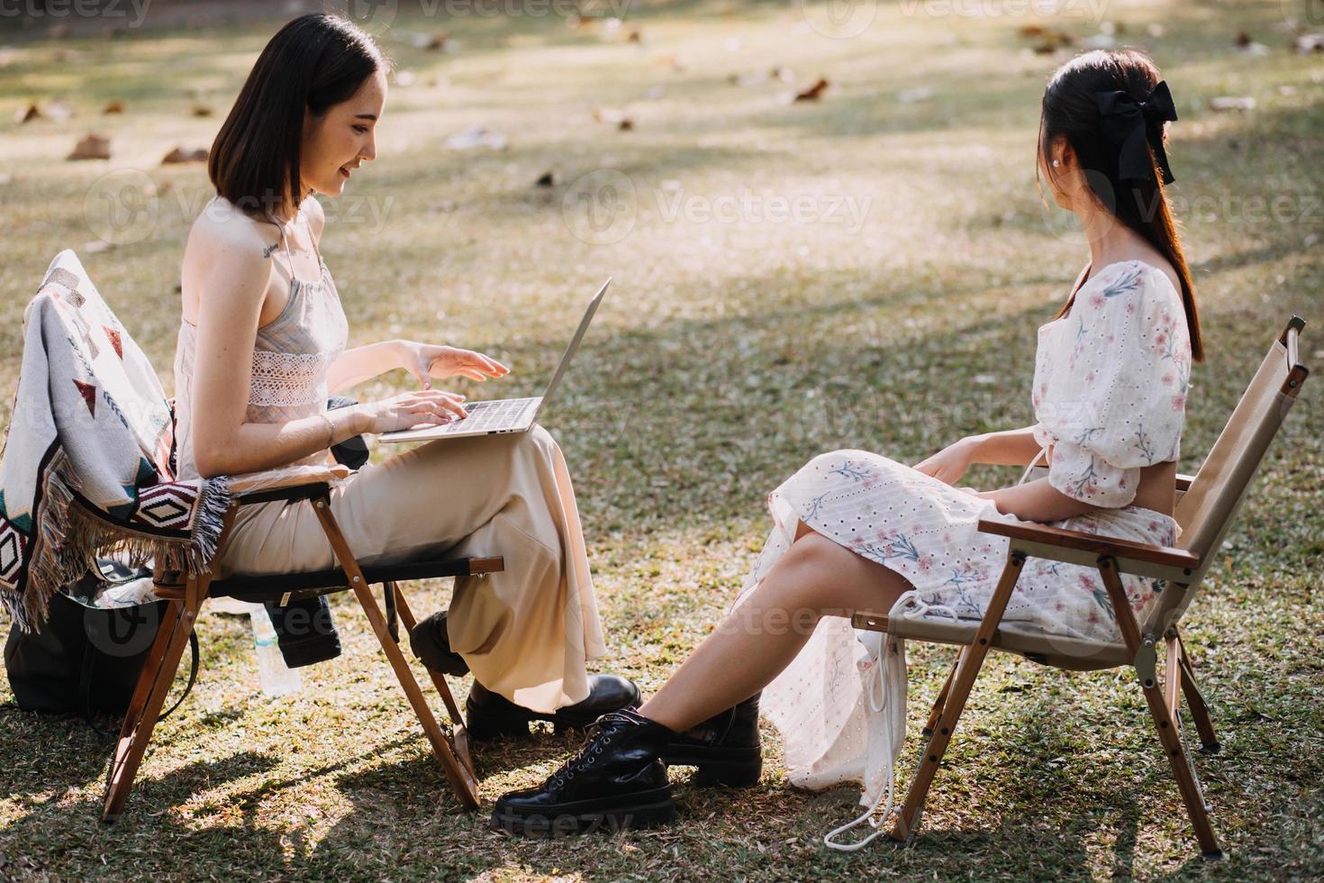 Young pretty brunette twin girls sitting on the grass with legs slightly bent in knees and looking in a brown book, wearing casual coat in autumn sunny weather on blurry background. photo