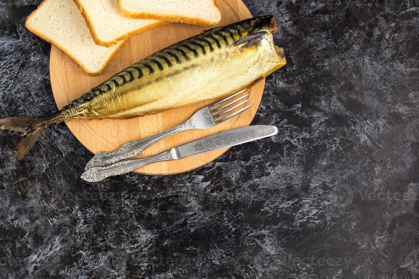 Smoked mackerel without head with fork knife cutting board bread on black background. copyspace top view photo