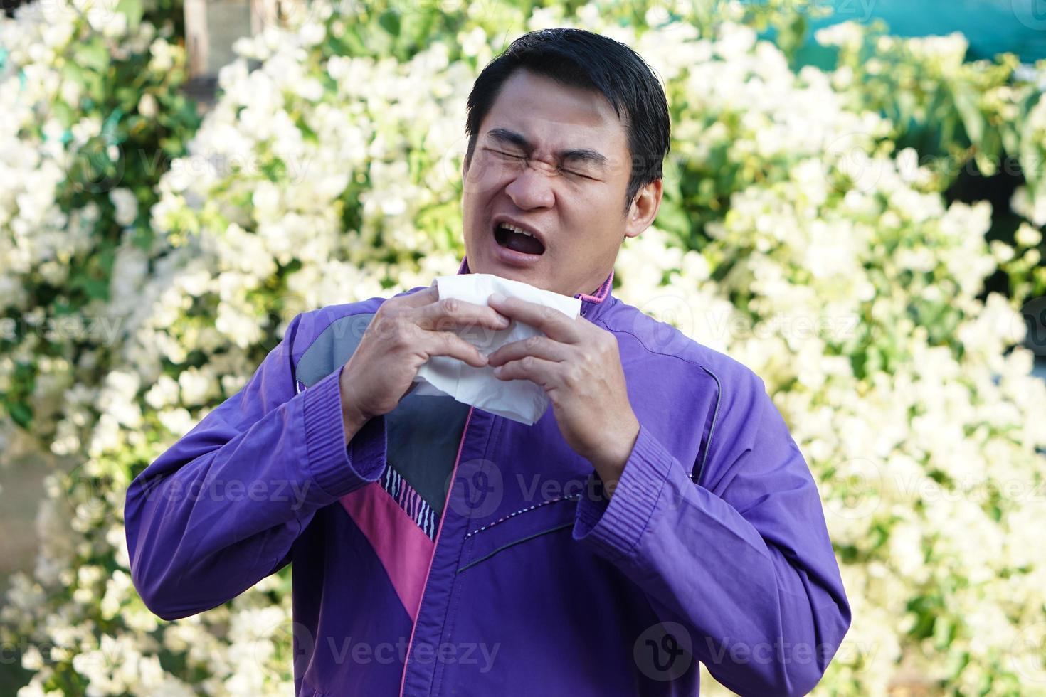 Asian man is sneezing, holding white tissue paper , open his mouth, standing beside blooming flowers at the park. Concept , Pollen allergy symptoms, sick, runny nose, catch a cold. health problems. photo