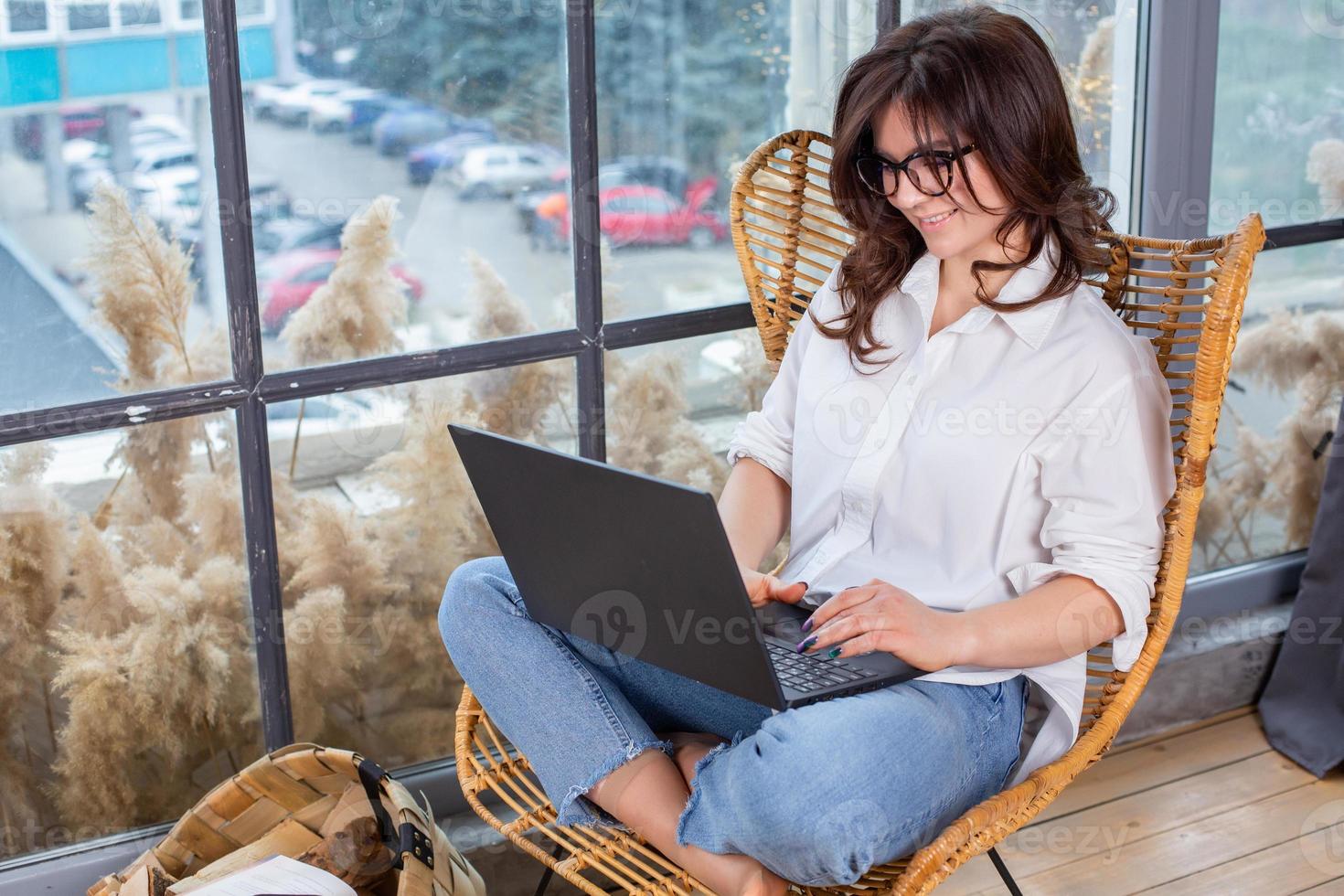 bella mujer de negocios trabajando en una laptop en una silla cerca de la ventana. mujer independiente que se conecta a internet a través de la computadora. blogger o periodista escribiendo un nuevo artículo. foto