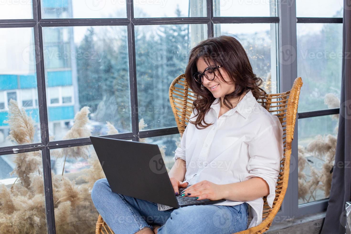 Beautiful businesswoman working on a laptop in a chair near the window. Female freelancer connecting to internet via computer. Blogger or journalist writing new article. photo