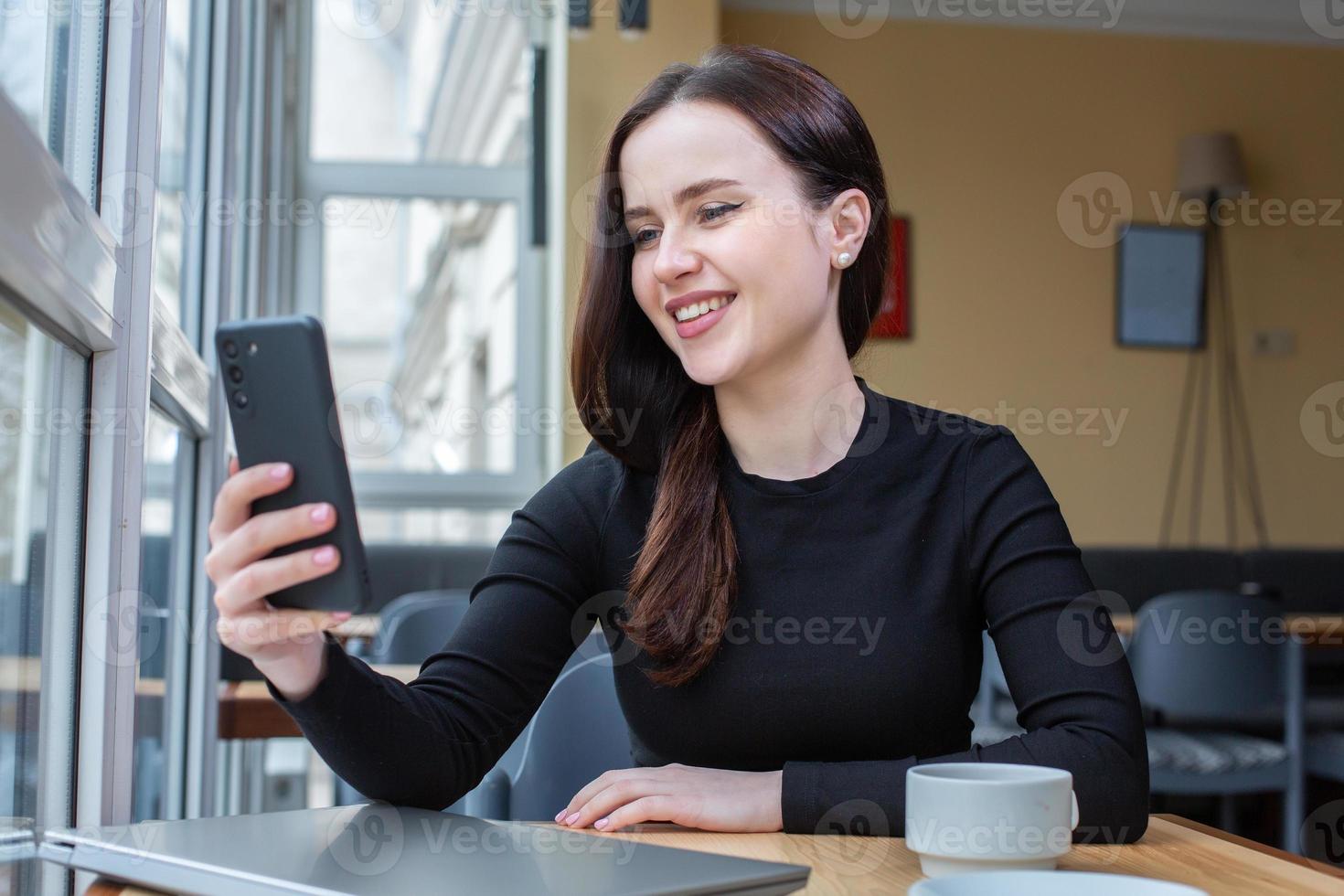 Portrait of smiling young woman using mobile phone while sitting in in coffee shop. White female freelancer connecting to internet on mobile phone. photo