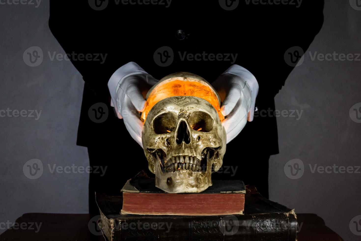 Portrait of Mysterious Figure Lifting the Top of a Human Skull on Stack of Ancient Books. Light After Death. Dark and Haunting. photo