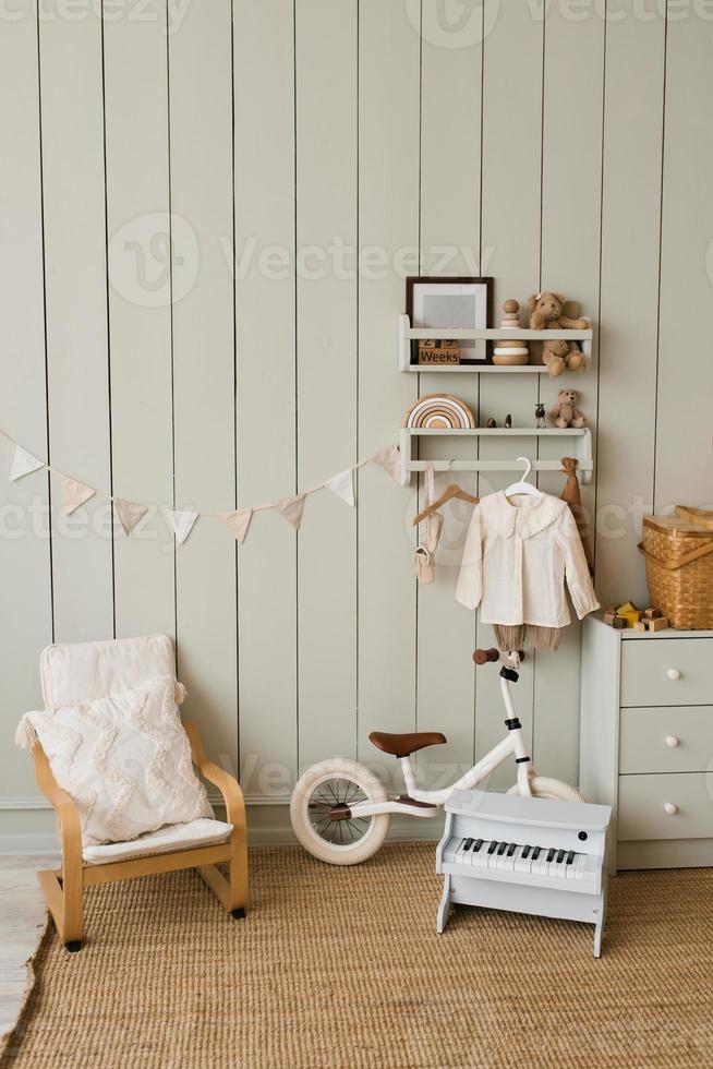 elegante composición de un acogedor interior escandinavo de una habitación infantil con sillón, juguetes de peluche y madera, una cesta de mimbre en la cómoda y banderas colgantes textiles foto