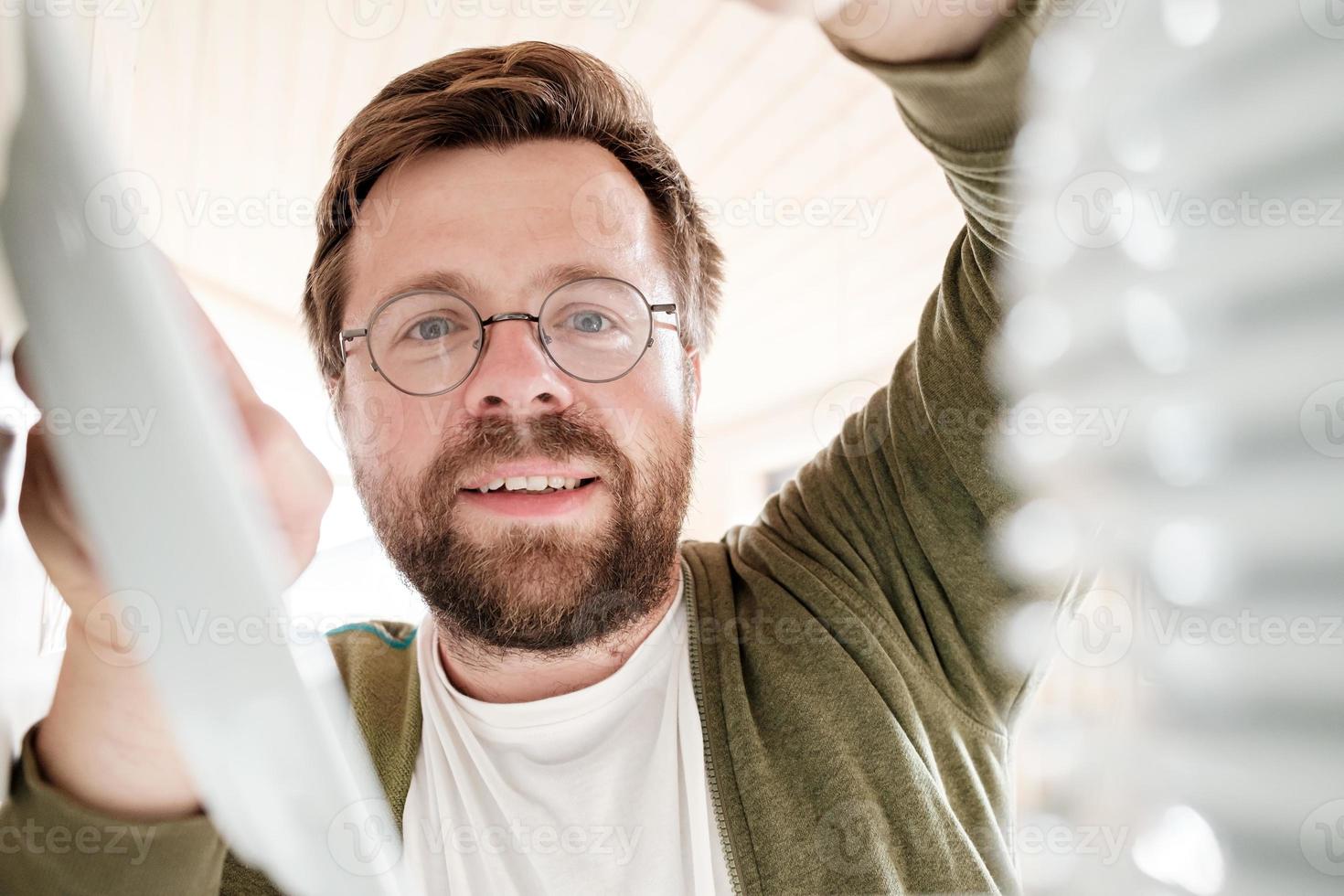 Camera view from the kitchen cabinet of a cute bearded man with glasses putting clean plates on the shelf. photo