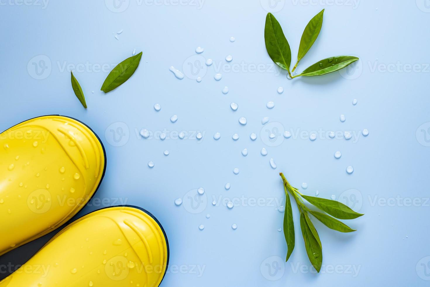 Yellow rubber boots stand next to a snowflake made of water drops and scattered green leaves, on a blue background. Top view. photo