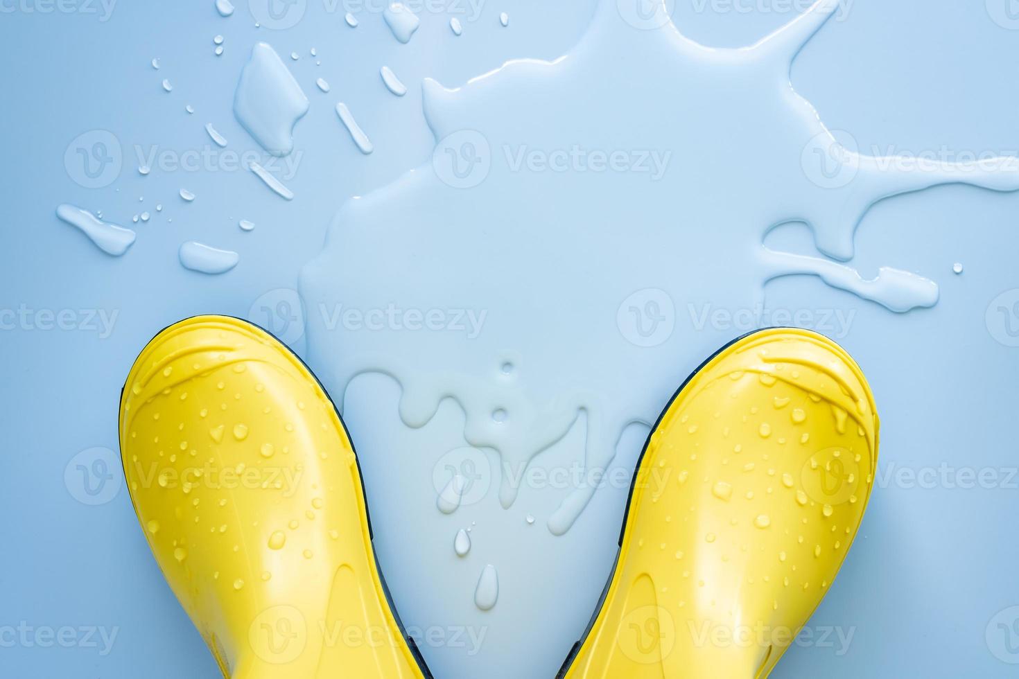 Yellow rubber boots stand near spilled water on a blue background. Concept of preparation for the rainy season. Copy space. photo