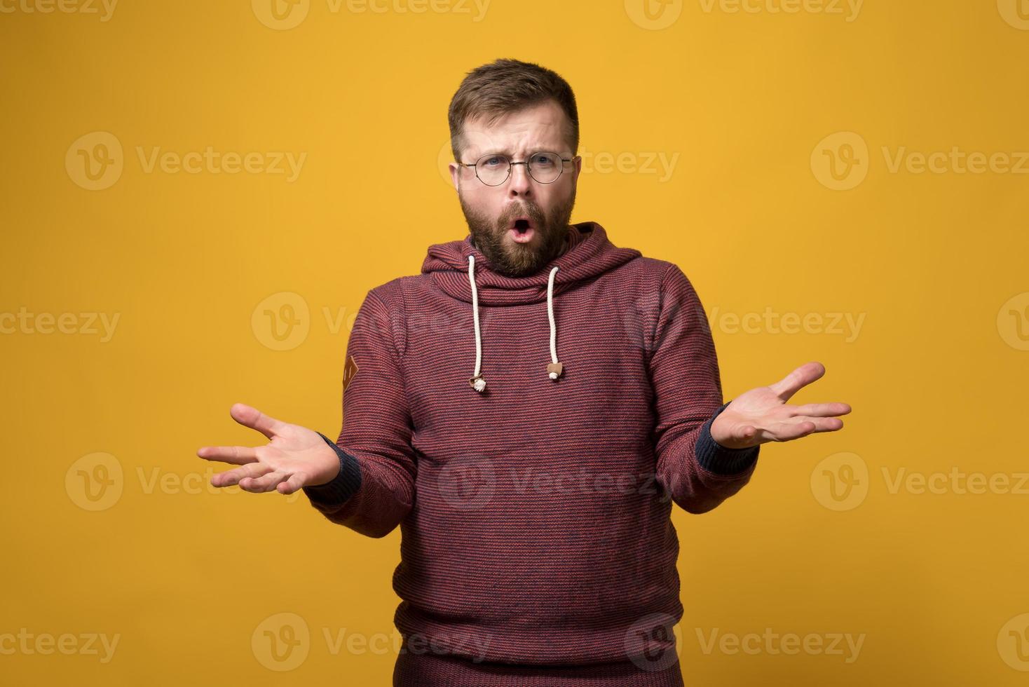 un hombre barbudo descontento y molesto con gafas levanta las manos desconcertado y dice algo mientras mira a la cámara. foto