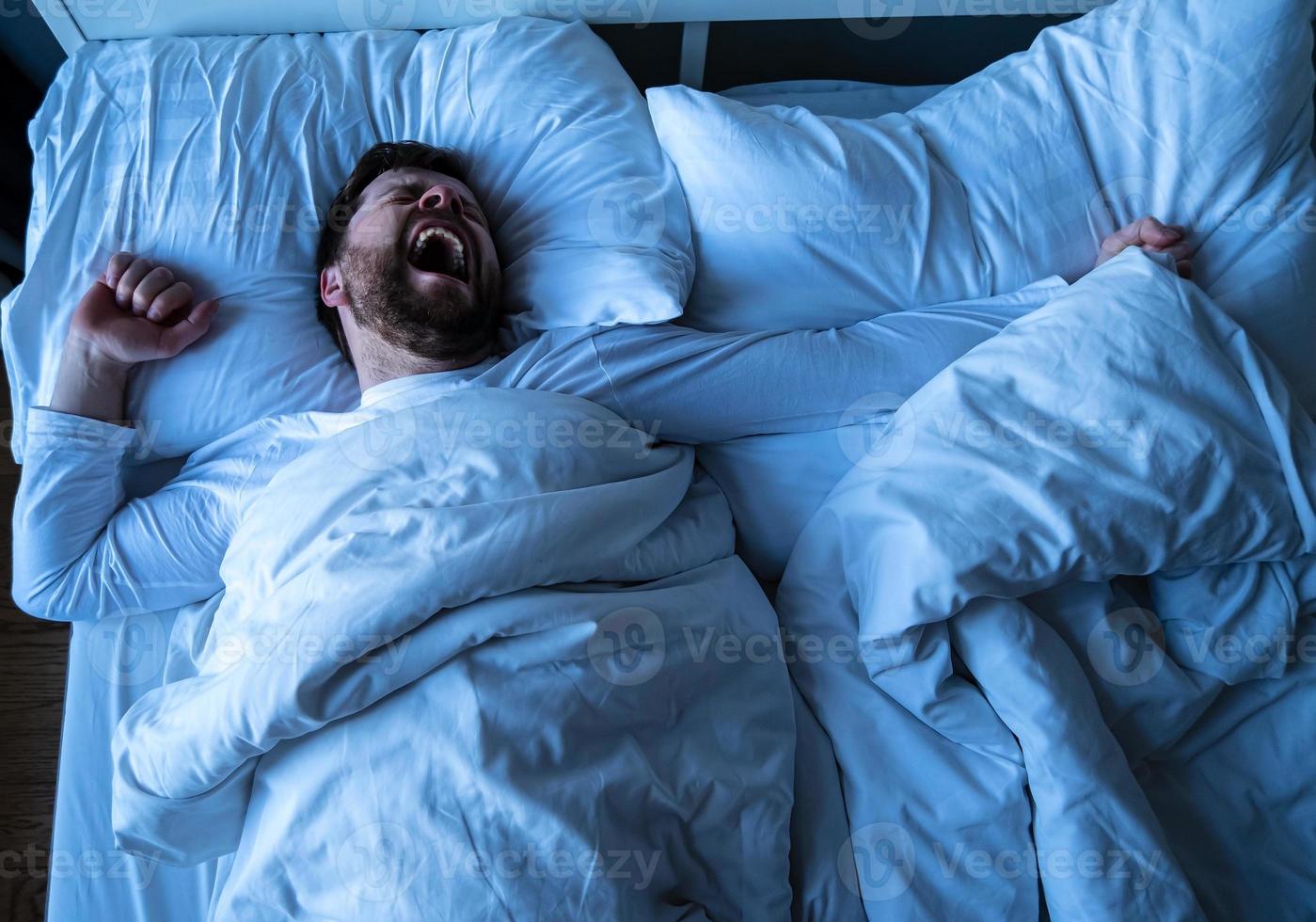 Young bearded man in white pajamas wakes up from sleep, he yawns and stretches as he lies in bed. photo