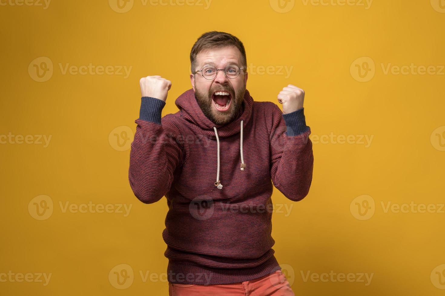 Happy Caucasian man makes a winner gesture, he energetically raises fists up and shouts in joy. Yellow background. photo