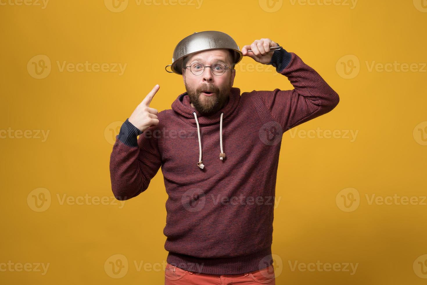 Pastafarianism. Cute man puts a colander on head and points at it with finger. Parody of religion. Studio, yellow background. photo