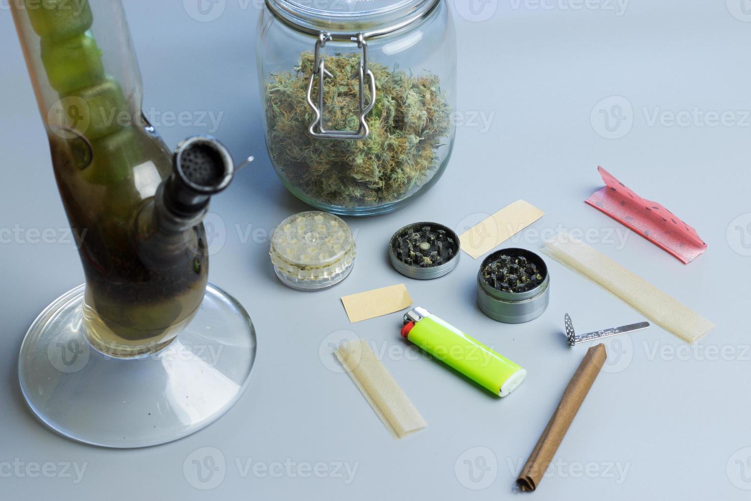 Smoking cannabis accessories and stuff on gray background. Bong, blunt and joint paper, grinder and marijuana buds in jar photo