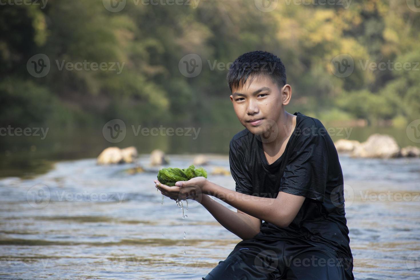 colegial asiático sosteniendo algas de agua dulce de bucear en el río y tirando de ellas para estudiar la fertilidad de la naturaleza del río y hacer el trabajo del proyecto escolar ambiental, en movimiento. foto